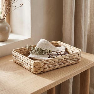 May include: A rectangular woven seagrass basket with a thick braided rim. Inside the basket rests a folded beige linen cloth, a small sprig of dried eucalyptus leaves, and a pair of brown tortoiseshell reading glasses with clear lenses.