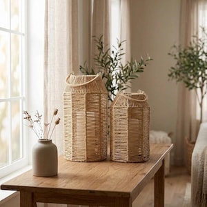 May include: Two hexagonal lanterns crafted from natural tan rope with integrated rope handles, positioned next to a smooth, light-colored ceramic vase containing dried botanical stems, all resting on a wooden surface.