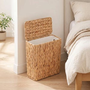 May include: A rectangular woven seagrass laundry hamper with a hinged lid and a white interior lining. The basket features a natural tan color and a thick braided texture, standing upright with an open lid beside a bed frame.
