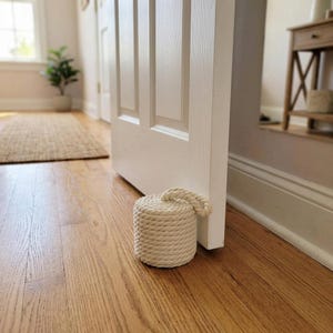 May include: A cylindrical doorstop crafted from thick, off-white braided rope. The object features a tight, woven pattern throughout its exterior and includes a small, integrated rope handle loop at the top for easy placement and movement.