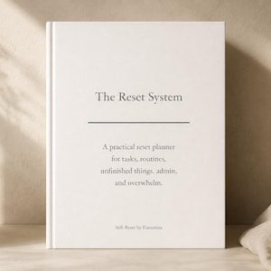 Może przedstawiać: Biała książka zatytułowana "The Reset System" z tekstem opisującym ją jako praktyczny planer resetowania. Książka leży na jasnej powierzchni, obok wazonu z suszonymi roślinami i złożonej tkaniny.