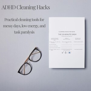 May include: An overhead shot features a white booklet with the text "ADHD Cleaning Hacks" and "The 15-Minute Dash." A teal teacup and saucer with gold trim, a pencil, and eyeglasses are also on the gray surface. The image suggests a theme of productivity and organization.