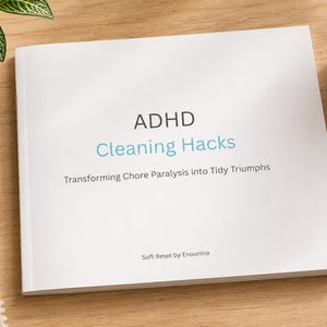 Puede incluir: Un libro blanco titulado "ADHD Cleaning Hacks" con el subtítulo "Transforming Chore Paralysis into Tidy Triumphs" está sobre una superficie de madera. Un bolígrafo y una taza de café están al lado del libro. También se ve una pequeña planta en maceta.