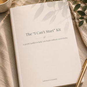 May include: A cream-coloured book titled "The 'I Can't Start' Kit" with the subtitle "A gentle toolkit to help you begin without overwhelm." A gold pen rests on the book. A mug of coffee and greenery are in the background.
