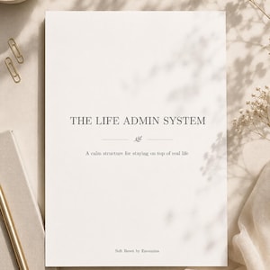 Może przedstawiać: Biały notatnik z tytułem "THE LIFE ADMIN SYSTEM" w kroju szeryfowym. Podtytuł brzmi "Spokojna struktura, aby być na bieżąco z prawdziwym życiem". Widoczny jest złoty długopis i spinacze do papieru.