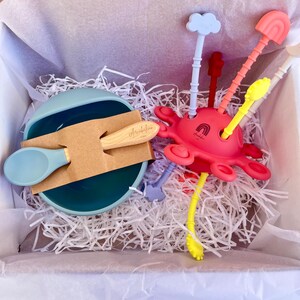 Può includere: Un set per l'alimentazione del bambino in una scatola bianca con riempimento di carta. Il set include una ciotola e un cucchiaio verde acqua, un cucchiaio di legno e un giocattolo a forma di polpo rosa con utensili colorati. Il giocattolo polpo ha un arcobaleno.