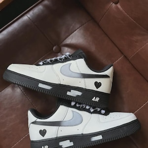 Pode incluir: Par de tênis brancos e pretos com logotipos swoosh cinza. Os sapatos têm solas pretas e a palavra "AIR" é impressa na lateral. Um coração preto está na lateral de cada sapato.