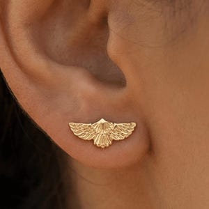 May include: A gold-colored earring in the shape of a stylized bird with outstretched wings. The earring is worn on the ear and is set against a blurred background.