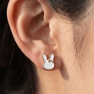 May include: A silver-toned bunny stud earring with a white enamel fill is shown on an ear. The bunny has long ears and a small bow. The earring is a small, delicate piece of jewelry.