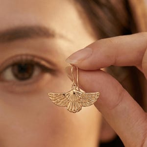 May include: A gold-colored earring with a wing-like design is held up in front of a person's eye. The earring has a detailed, textured surface, with the wing shape clearly defined. The earring's hook is visible, and the overall aesthetic is elegant.