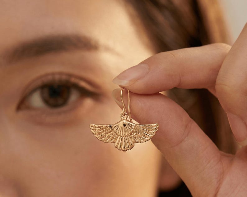 May include: A gold-colored earring with a wing-like design is held up in front of a person's eye. The earring has a detailed, textured surface, with the wing shape clearly defined. The earring's hook is visible, and the overall aesthetic is elegant.