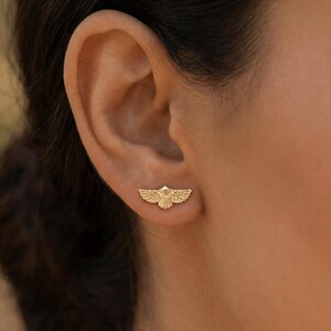 May include: A gold-colored earring in the shape of a stylized bird with outstretched wings. The earring is worn on the ear and is visible in a close-up shot. The earring has detailed feather patterns.