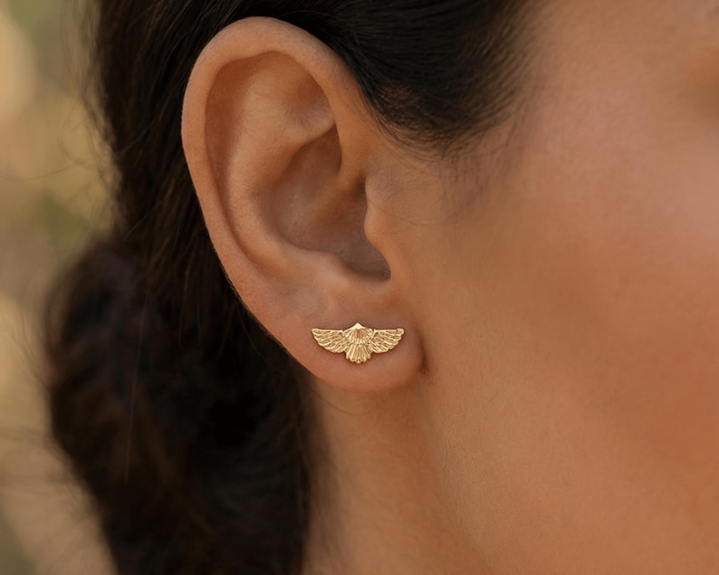 May include: A gold-colored earring in the shape of a stylized bird with outstretched wings. The earring is worn on the ear and is visible in a close-up shot. The earring has detailed feather patterns.
