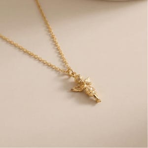 May include: A gold-colored necklace featuring a small cherub pendant. The cherub has detailed wings and is suspended from a delicate chain. The necklace is displayed on a neutral background, highlighting the jewelry's design and color.