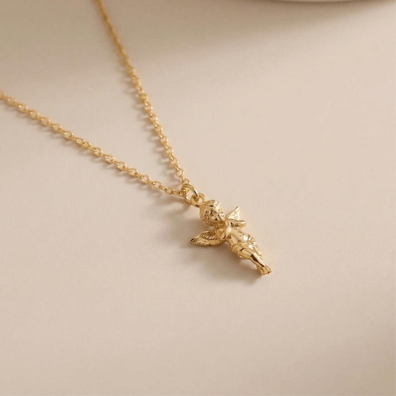 May include: A gold-colored necklace featuring a small cherub pendant. The cherub has detailed wings and is suspended from a delicate chain. The necklace is displayed on a neutral background, highlighting the jewelry's design and color.