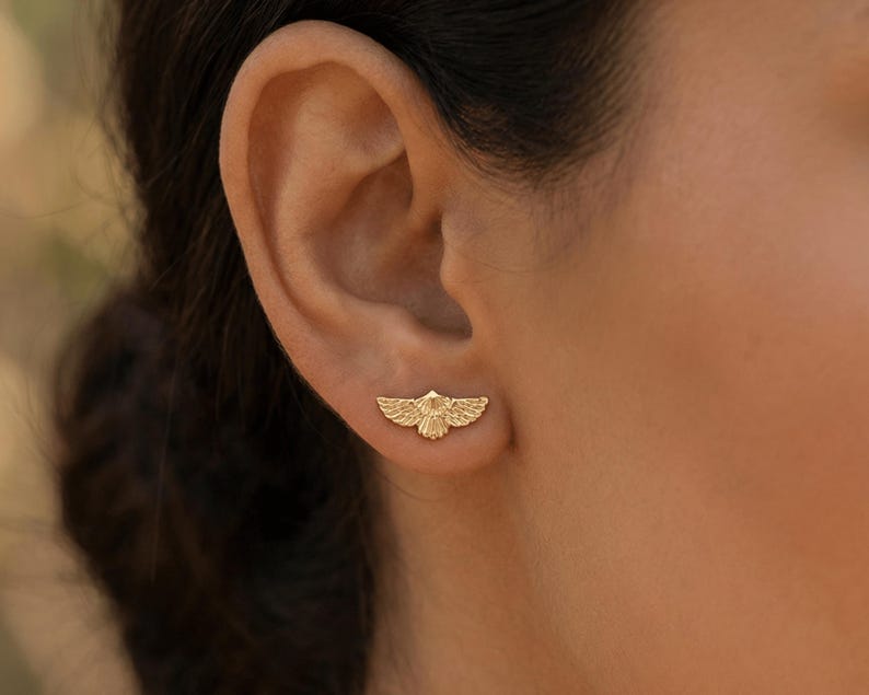 May include: A gold-colored earring in the shape of a stylized bird with outstretched wings. The earring is worn on the ear and is set against a blurred background.