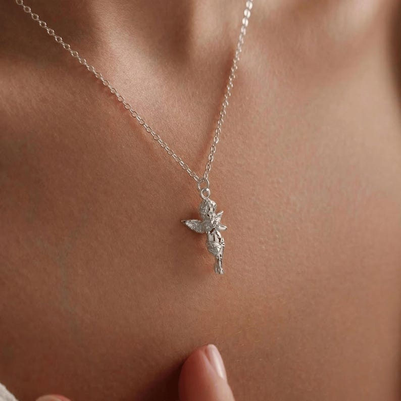 May include: A silver-colored necklace featuring a small cherub pendant. The cherub has detailed wings and is suspended from a delicate chain. The necklace is shown against a skin-toned background, highlighting the jewelry's design and craftsmanship.