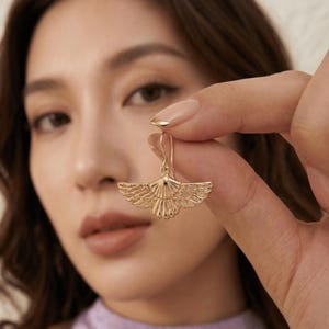 May include: A gold-colored earring with a stylized wing design is held up. The earring has a detailed, textured surface. The earring is a drop style, with a hook at the top. The background is blurred, with a person's face and hand visible.