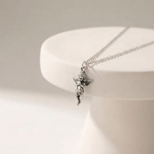 May include: A silver-colored necklace featuring a cherub pendant with detailed wings and clasped hands. The cherub is suspended from a delicate chain. The necklace is displayed on a white, rounded surface.