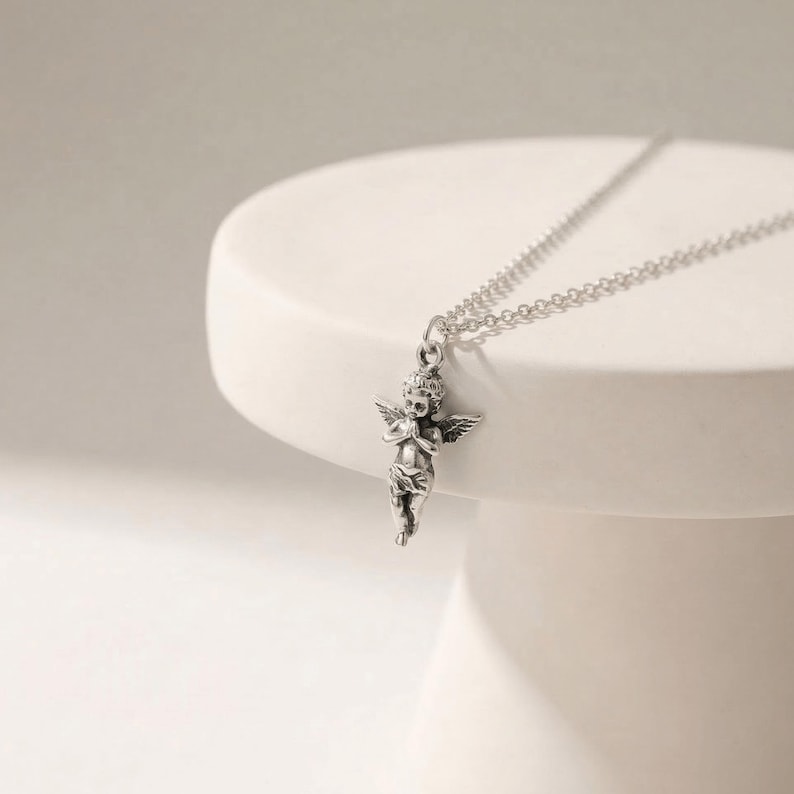 May include: A silver-colored necklace featuring a cherub pendant with detailed wings and clasped hands. The cherub is suspended from a delicate chain. The necklace is displayed on a white, rounded surface.
