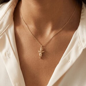 May include: A gold-colored necklace featuring a small cherub pendant. The cherub has outstretched wings and is suspended from a delicate chain. The necklace is worn against a white collared shirt.