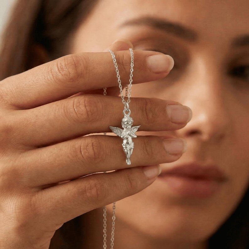 May include: A silver necklace featuring a detailed cherub pendant with outstretched wings. The cherub is in a praying pose, suspended from a delicate chain. The necklace is held up against a blurred background.