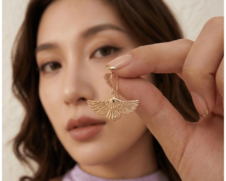May include: A gold-colored earring with a stylized wing design is held up. The earring has a detailed, textured surface. The earring is a drop style, with a hook at the top. The background is blurred, with a person's face and hand visible.