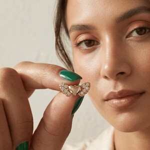 K&ouml;nnte beinhalten: Ein goldfarbener herzf&ouml;rmiger Ring mit Diamantakzenten. Der Ring hat eine zentrale Herzform, die von zwei fl&uuml;gelartigen Designs flankiert wird, die jeweils mit kleinen, funkelnden Diamanten besetzt sind. Der Ring wird vor einem neutralen Hintergrund gehalten.