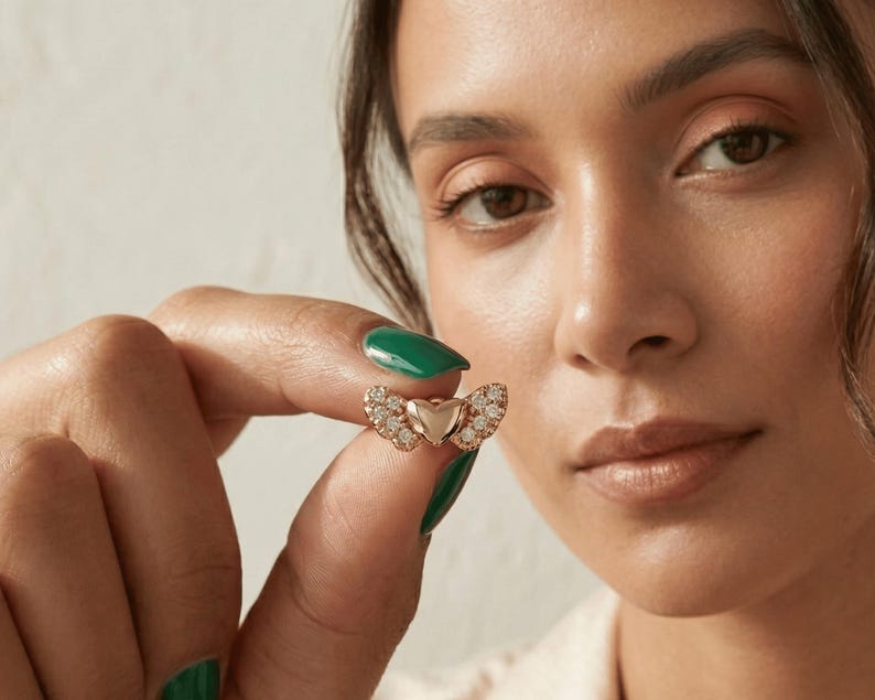K&ouml;nnte beinhalten: Ein goldfarbener herzf&ouml;rmiger Ring mit Diamantakzenten. Der Ring hat eine zentrale Herzform, die von zwei fl&uuml;gelartigen Designs flankiert wird, die jeweils mit kleinen, funkelnden Diamanten besetzt sind. Der Ring wird vor einem neutralen Hintergrund gehalten.
