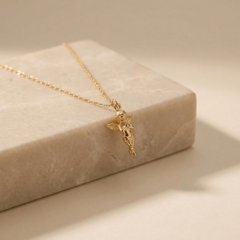 May include: A gold-colored necklace featuring a small cherub pendant. The cherub has detailed wings and is suspended from a delicate chain. The necklace is displayed on a light-colored, rectangular surface, creating a minimalist aesthetic.