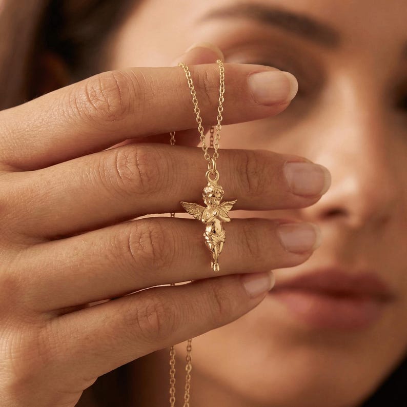 May include: A gold-colored necklace featuring a cherub pendant. The cherub has detailed wings and is in a praying pose. The chain is delicate and the pendant is suspended from it. The necklace is held up against a blurred background.