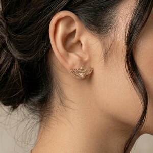 May include: A close-up of a person's ear with a gold-colored stud earring. The earring is in the shape of a stylized bird with outstretched wings. The earring is small and delicate, and the details of the bird are visible.