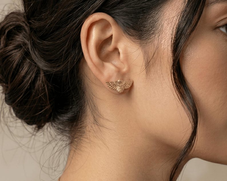 May include: A close-up of a person's ear with a gold-colored stud earring. The earring is in the shape of a stylized bird with outstretched wings. The earring is small and delicate, and the details of the bird are visible.