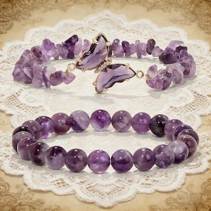 May include: Two amethyst bracelets. One features a butterfly-shaped clasp with purple gemstone wings and gold-colored metal. The other bracelet is made of round, polished amethyst beads. Both bracelets are displayed on a white lace surface.