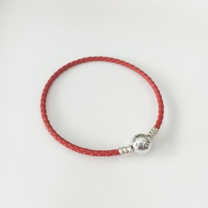 Puede incluir: Una pulsera de cuero trenzado rojo con un cierre redondo de metal plateado. El cierre es esférico con una superficie lisa y texto grabado, conectado al cordón trenzado por dos tapas de extremo de metal plateado estriado a cada lado.
