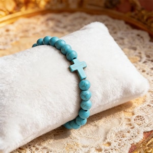May include: A turquoise beaded bracelet with a cross charm. The bracelet is made of round, light blue beads and features a matching cross. The bracelet is displayed on a white, fluffy surface.