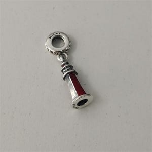 Puede incluir: Un charm de plata con forma de faro y un detalle de esmalte rojo. El anillo superior está grabado con el texto S925 ALE, y la base del faro también presenta la inscripción grabada S925 ALE. El charm tiene un acabado metálico.