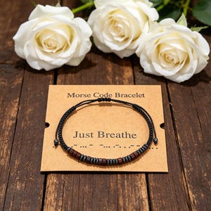 Könnte beinhalten: Ein schwarzes Schnurarmband mit abwechselnden dunkelbraunen Holzperlen und kleinen metallisch grauen Perlen in einem Morsecode-Muster. Das Armband liegt auf einer beigen Karte mit dem Text Morse Code Bracelet und Just Breathe.