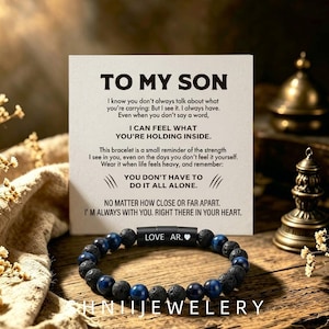 May include: A beaded bracelet featuring alternating dark blue and porous black beads with a central black cylindrical clasp engraved with the text LOVE AR. and a heart symbol, displayed in front of a white card with the printed text TO MY SON.
