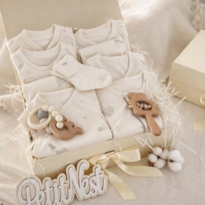 Può includere: Una scatola regalo color crema piena di vestiti per bambini, calzini e giocattoli di legno. La scatola è legata con un nastro color crema e include un cartello di legno con la scritta "Petit Nest".