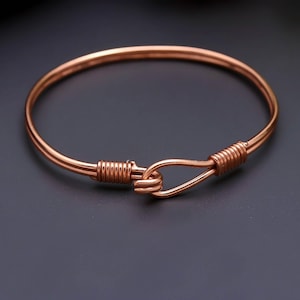 Può includere: Un bracciale rigido in metallo color rame con un meccanismo di chiusura a gancio. Il bracciale è realizzato con un filo metallico continuo che forma un anello circolare, con entrambe le estremità avvolte in spirali strette per la chiusura.