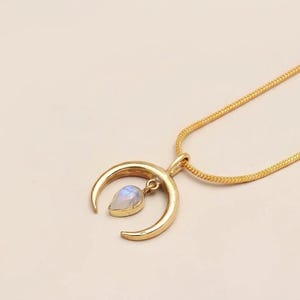 Può includere: Una collana color oro con un ciondolo a forma di mezzaluna dalla finitura liscia e lucida. Sospesa al centro della mezzaluna si trova una piccola pietra di luna a forma di goccia con un riflesso traslucido blu e bianco.