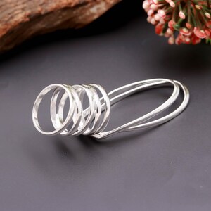 Könnte beinhalten: Ein silberfarbener Mehrfingerring, der aus einer Reihe von fünf miteinander verbundenen, horizontal ausgerichteten kreisförmigen Bändern besteht. Die Struktur ist aus einem durchgehenden, polierten Metalldraht gefertigt und bildet eine schlanke, längliche Form.