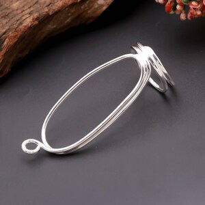 May include: A silver-coloured metal jewellery component featuring a large, elongated oval frame with a smooth, reflective surface. One end of the oval connects to a small, circular jump ring, while the opposite end features two curved, parallel bands.
