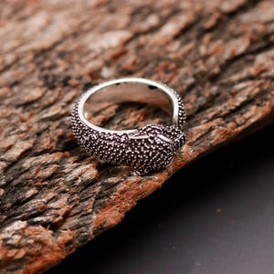 May include: A silver ring with a textured, dark-colored design. The ring features a detailed, possibly animal-inspired, head design. The ring is set against a natural, textured wooden surface.