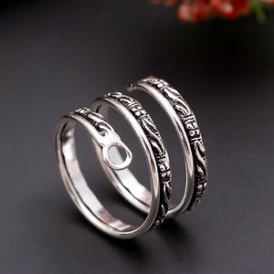 May include: A silver-toned metal ring featuring a bypass spiral design that wraps around the finger. The band has an oxidized finish with intricate embossed floral and geometric patterns along the outer surface and a small circular loop end.