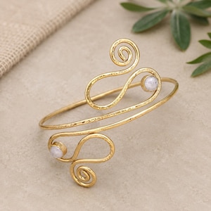 Puede incluir: Un brazalete dorado con un diseño en espiral y dos pequeñas piedras blancas redondas. El brazalete tiene una textura martillada y un detalle en espiral en la parte superior e inferior. La joya se exhibe sobre una superficie neutra.