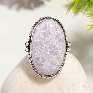 Pode incluir: Anel oval de prata com uma pedra roxa clara. A pedra tem um padrão floral e está definida em uma banda de prata detalhada. O anel é exibido em uma superfície branca.