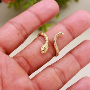 May include: A gold-colored snake ring wraps around a finger. The ring features a detailed snake head and body, with textured scales. The snake's head is positioned near the top of the finger, and the body curves around the finger.