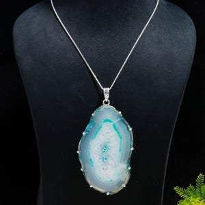 May include: A large, pendant necklace featuring a polished, blue and white agate stone. The stone is encased in a silver-colored metal setting and hangs from a delicate silver chain. The agate has a unique, natural pattern.